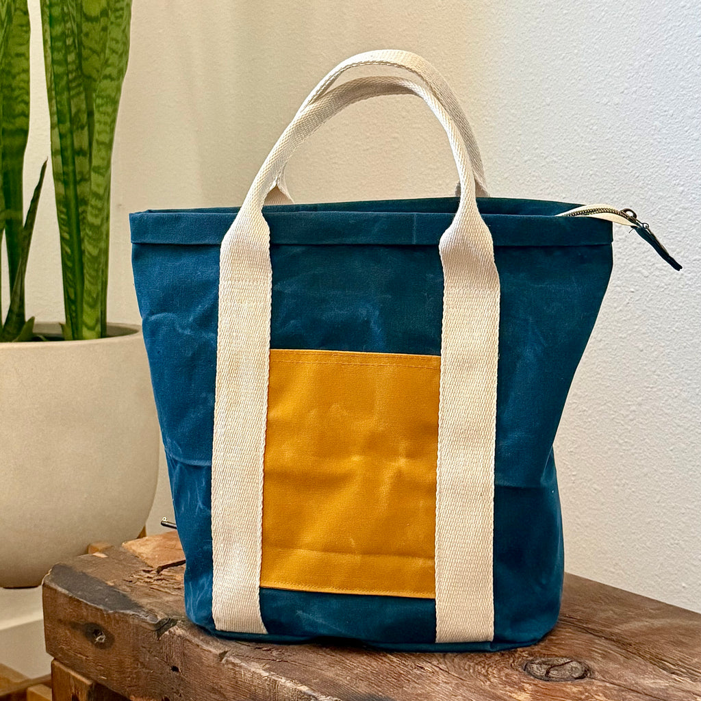 Blue and mustard tote bag with white handles on a wooden surface next to a plant.
